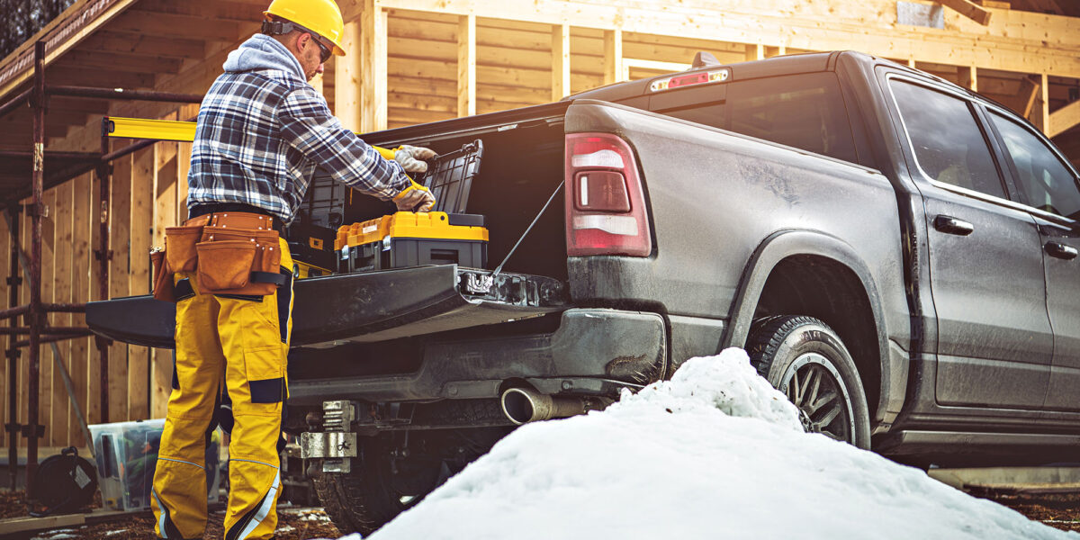 A construction worker in yellow gear prepares tools from a truck at a snowy building site. Wooden frames are visible, indicating an active construction project.