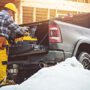 A construction worker in yellow gear prepares tools from a truck at a snowy building site. Wooden frames are visible, indicating an active construction project.