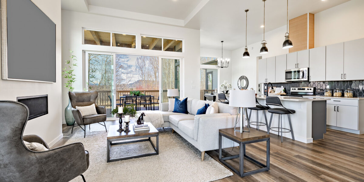 open concept living room with stylish furniture electric fireplace grey leather arm chairs windows revealing blue sky hardwood floors and view to kitchen