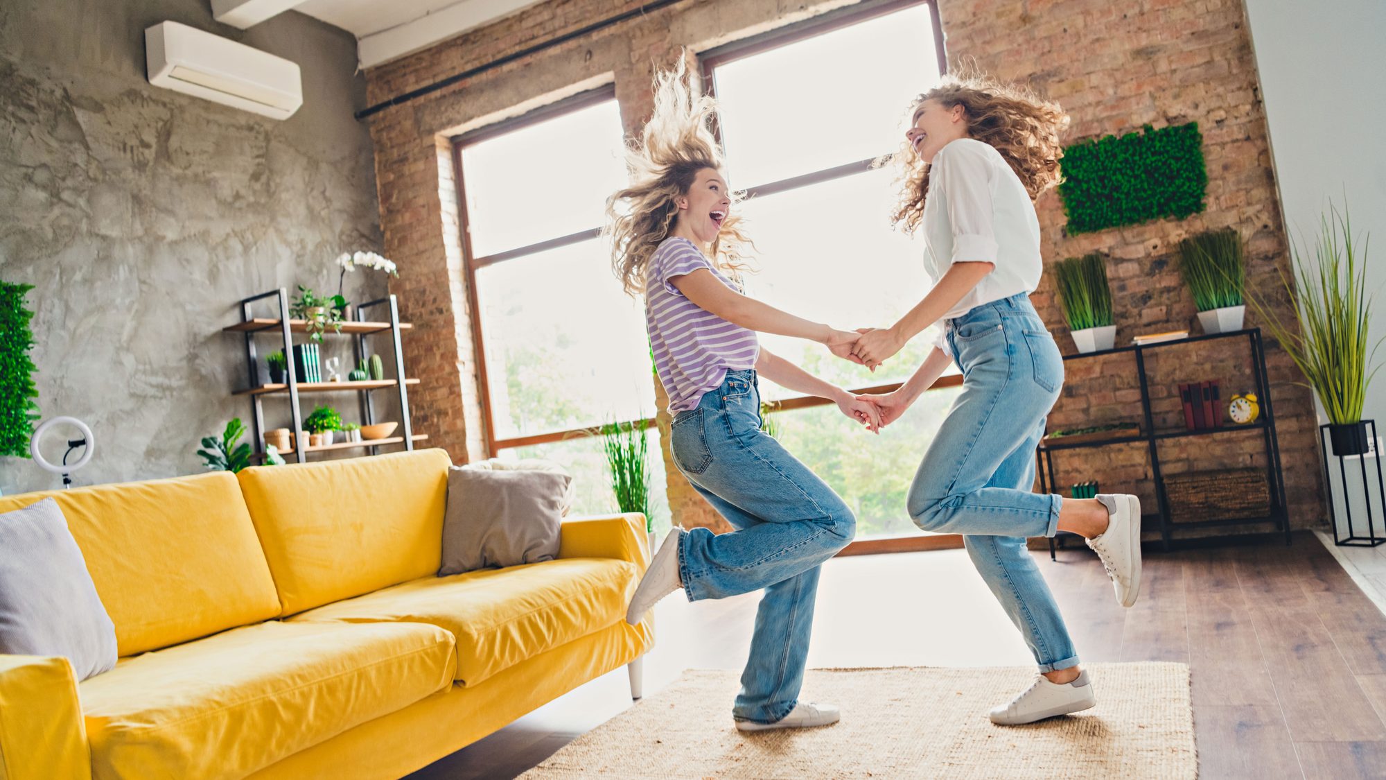 Two joyful women dancing in a stylish living room with a yellow sofa